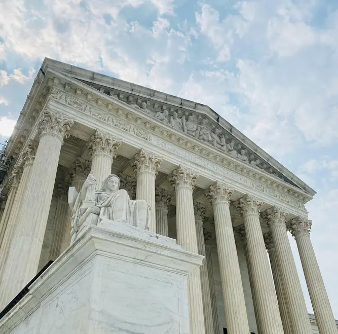 A low-angle view of the U.S. Supreme Court building in Washington D.C., featuring the front facade with Corinthian columns and a carving of a seated figure in the foreground.