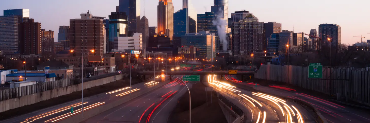 City skyline at dusk with blurred lights from vehicles on the highway in the foreground and tall buildings in the background.