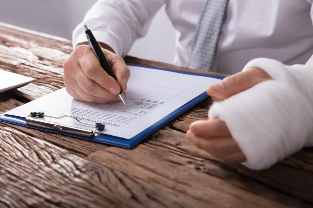 A person with a bandaged hand is filling out a form on a clipboard using a pen. The person is wearing a white shirt and a gray tie.